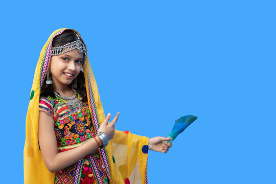 Portrait of happy indian girl kid wearing traditional colorful outfit holding painting brush against blue background, interior of new house wall, studio shot. copy space.
