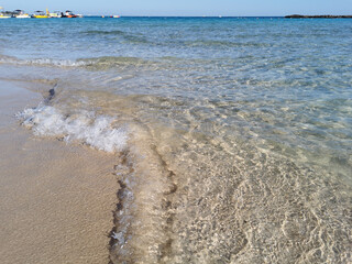 A small wave runs on the sandy shore of the beach in Fig Tree Bay against the background of the clear water of the Mediterranean Sea and the blue cloudless sky.