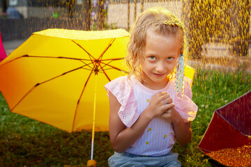 Cute little blonde girl with umbrella under rain drops on lawn in a sunny summer day