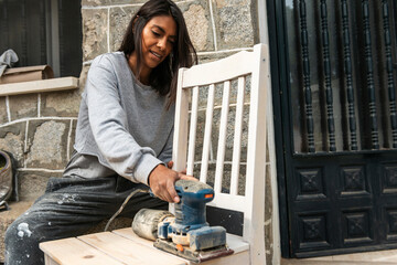 Brown skinned woman using sanding machine to restore a chair at home