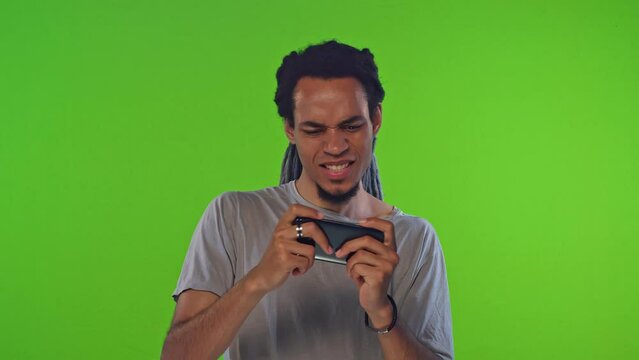 Young emotional ethnic African American man gamer playing video game G-Sensor controlling smart phone competing in online race with accelerometer experiencing excitement stands on green background