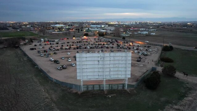 Outdoor drive-in movie theater at dusk. Drone shot rising up behind screen for aerial view of cars parked in dirt lot.