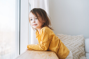 Cute little toddler girl in yellow longsleeve looking in window at home