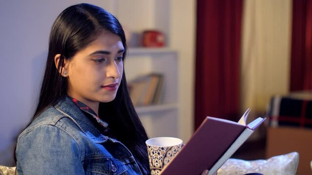 A college student wearing reading an interesting book at-home - education and learning  a favorite pastime  literature. A beautiful girl with black hair drinking a cup of hot tea / coffee while rel...