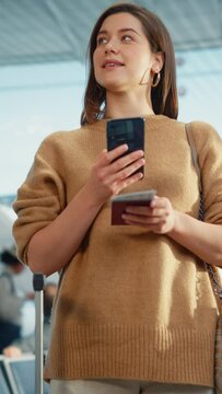 Vertical Screen. Airport Terminal: Happy Traveling Caucasian Woman Waiting at Flight Gates for Plane Boarding, Uses Mobile Smartphone, Checking Trip Destination on Internet. Smiling Female on a Trip