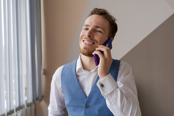 Pleasant handsome bearded man in shirt and waistcoat talking on phone, ordering taxi or food delivery to office at work. Smiling young caucasian businessman holding a call with partners clients.