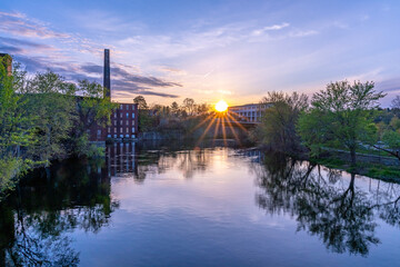 The sun and its rays at sunset with reflection on the Nashua River. On the shore there is a historical building of a cotton factory with a tall brick chimney in an old industrial park. Nashua, NH