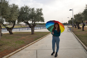 Young woman, red hair, freckles, with a rainbow umbrella, happy, in an outdoor park. Concept color, happiness, well-being, fun, rain.
