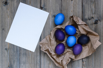 Top view flat lay on a rustic wooden table on which a white empty sheet of paper and blue and purple painted Easter eggs on craft paper are placed, copy space.