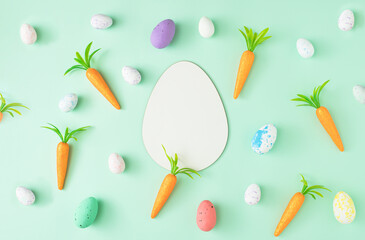Easter eggs and carrots arranged on a pastel green background with an egg shaped card note. Holiday flat lay minimal composition.