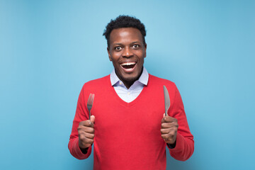 Hungry african young man holding fork and knife on hand ready to eat