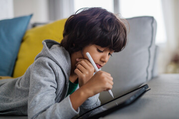 Little multiracial boy with tablet lying on sofa at home.