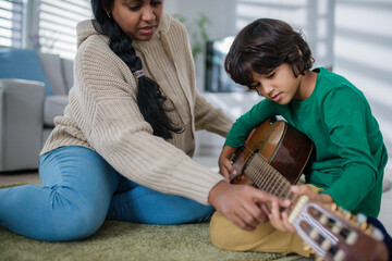 Little multiracial boy learning to play the guitar with his mother at home.