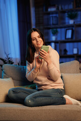 Obraz premium Young beautiful woman using mobile phone apps, browsing internet, social media, looking at smartphone and smiling. Home leisure in the evening, cinematic lighting.