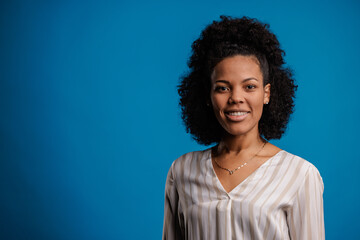 Lovely african-american woman in casual outfit and curls, smiling, on blue background.