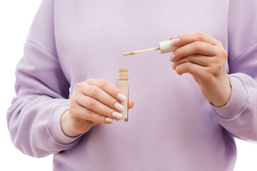 Close up shot of a woman in a lavander oversized sweatshirt holding a concealer bottle and a brush in hands. Nice groomed nails with nail polish.