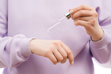 Close up shot of a woman in a lavander oversized sweatshirt holding a pipette with serum in hands. Serum drop is hanging. Nice groomed nails with nail polish.