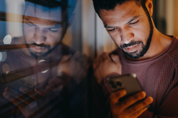 Young man with a smart phone looks sad, standing by window in evening at home.