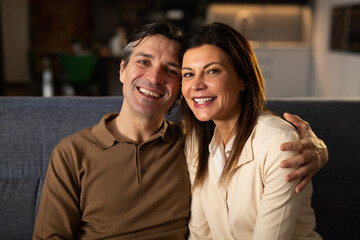 Cinematic shot of young happy married couple is smiling in camera while relaxing and enjoying rest together by leaning on soft comfortable sofa in living room at home.