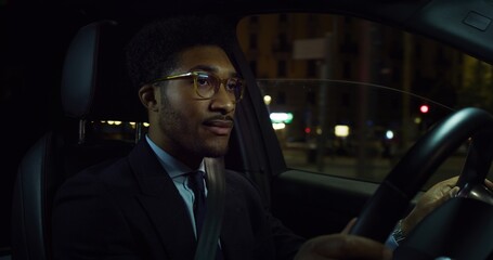 Cinematic shot of young handsome afro american successful elegantly dressed businessman with glasses is driving modern luxury car in center of the city by night.
