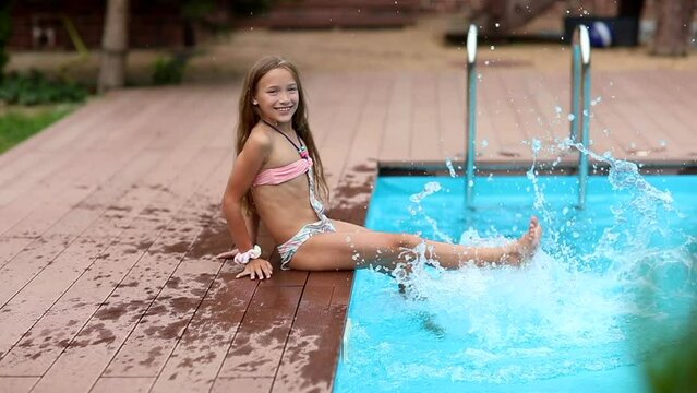 Beautiful long haired girl in bikini splashing water near blue swimming pool at the back yard. Summertime. Slow motion.