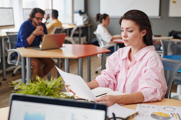 Fototapeta premium Concentrated young Caucasian businesswoman sitting in coworking space and working with paper