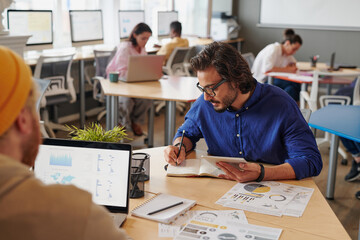 Concentrated young mixed race man in glasses sitting at table and making notes in organizer while planning new project