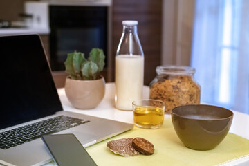 Table set for breakfast, kitchen in the background. Natural meal and technology devices. Interior view of a cozy and modern home. Millennial, youth, home working, home schooling, remote work concept.