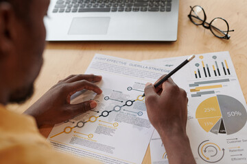 Over shoulder view of black expert sitting at table and analyzing effectivieness of business