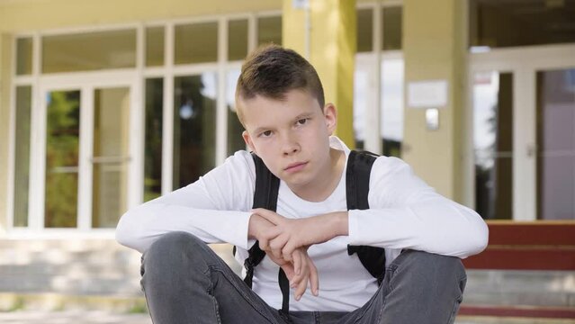 A Caucasian teenage boy looks seriously at the camera as he sits in front of school - closeup