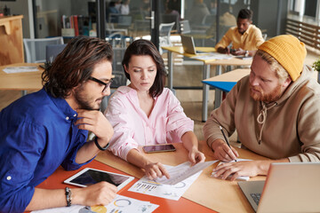 Attractive young woman pointing at paper data while analyzing chart with colleagues in shared workplace
