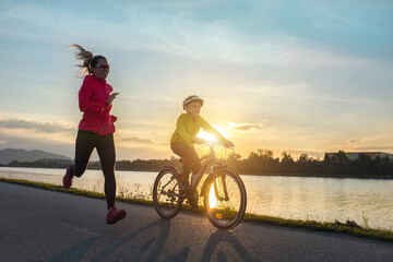 Happy mother and son go in sports outdoors. Boy rides bike in helmets, mom runs on sunny day. Silhouette family at sunset. Fresh air. Health care, authenticity, sense of balance and calmness