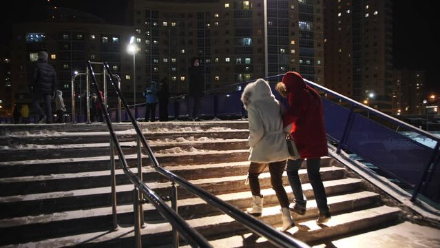 Man and woman in warm coats go upstairs on snowy street at winter night back view slow motion. City life and modern architecture
