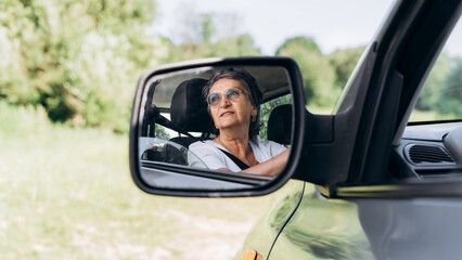Senior cheerful smiling female driver behind wheel of car looking away, reflection in side rearview mirror outdoors. Caucasian elderly woman driving car, close-up