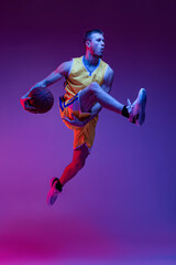 Studio shot of muscled man, basketball player training with ball isolated on purple background in...