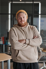 Portrait of smiling young bearded Caucasian man in hat and hoodie standing with crossed arms in office