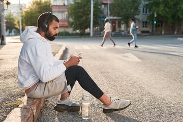 Athletic young middle eastern man in wireless headphones texting message on phone while relaxing on curb in city