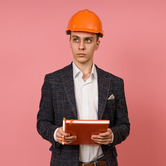 young man engineer builder in a hard hat with a book in his hands on a pink background