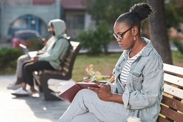 Obraz premium Concentrated young African-American woman in denim outfit and making notes in organizer while working on project in city