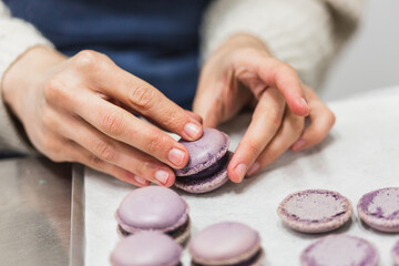 Hands of pastry chef joining lids of purple macaron with filling on table