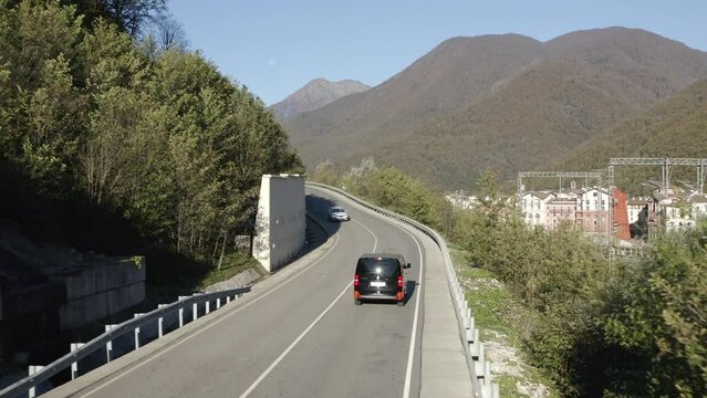 Black minivan car riding on automobile traffic asphalt mountain road amazing panorama sunny luxury resort village cityscape. Aerial view automobile bus ride on serpentine path mountain