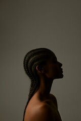 Vertical side portrait of African young woman with braids posing on dark wall.