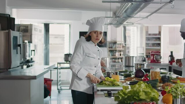 Portrait of female cook cutting green celery in gourmet kitchen, using cutting board and knife to slice coleslaw salad. Woman chef with uniform preparing ingredients for food recipe.