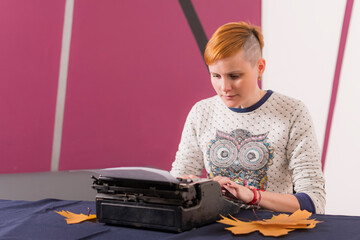 Serious young girl with red hair and stylish hairstyle sits at table and types text on old typewriter.