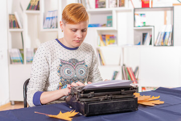 Serious young girl with red hair and stylish hairstyle sits at table and types text on old typewriter.