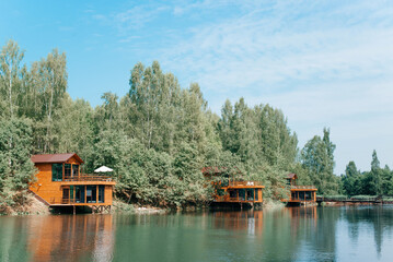 Exterior of modern country holiday complex on lake, wooden two-story houses on water near forest on sunny summer day. Aesthetic villas with balcony, outdoors by lake