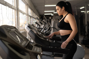Naklejka na ściany i meble Cardio workout. Side view of happy athletic woman in young sportswear adjusting speed on treadmill while exercising in gym or sport club.