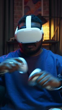 Vertical Portrait: Young Stylish Black Man Putting On Virtual Reality Headset. African American Man Sitting on a Sofa at Home in the Evening, Playing VR Video Games. Dramatic Zoom In Shot.