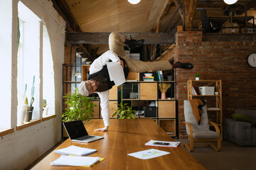 Young bearded man, office clerk having fun, doing yoga on wooden table in modern office at work time with gadgets. Concept of business, healthy lifestyle, sport, hobby
