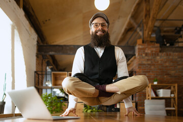 Emotional office clerk, young man doing yoga on wooden table in modern office at work time with...